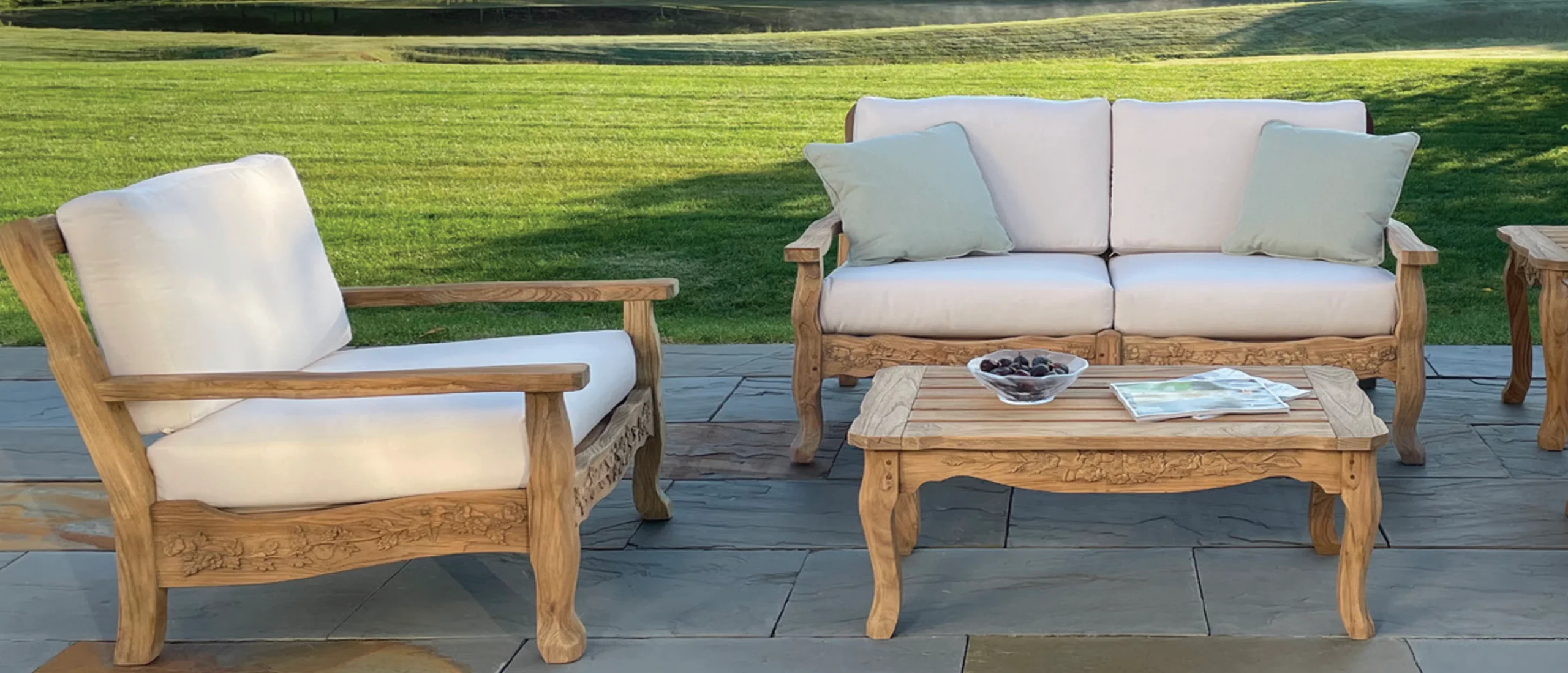 Carved teak outdoor lounge chair, sofa and coffee table with cream cushions on a bluestone patio overlooking a lawn