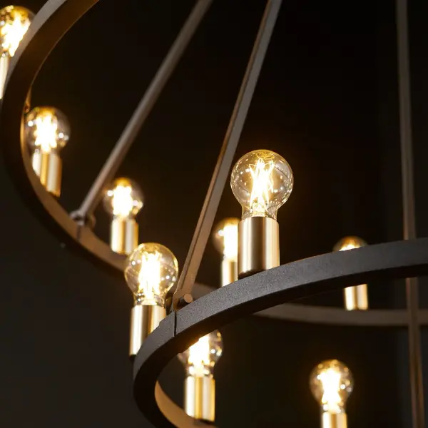 The Paxton wagon wheel chandelier makes a dramatic impact with 24 lights arranged in a geometric conical design. Two-tone noir and aged brass finishes create visual interest while stone seeded glass shades add rustic texture. Damp-rated for versatility. - alternate view