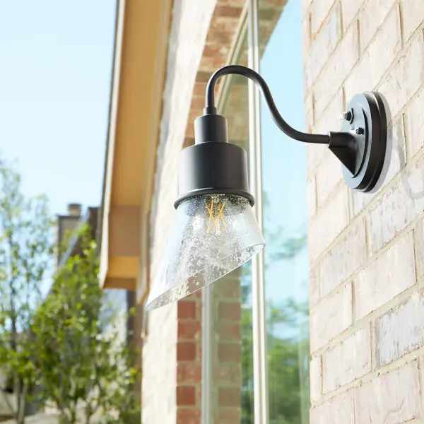 The Torrey wall light combines industrial and rustic elements with a softly curved arm and hood shade. Its Wet rating makes it perfect for outdoor use, while the clear seeded glass and noir finish create a versatile look suitable for various settings. - alternate view