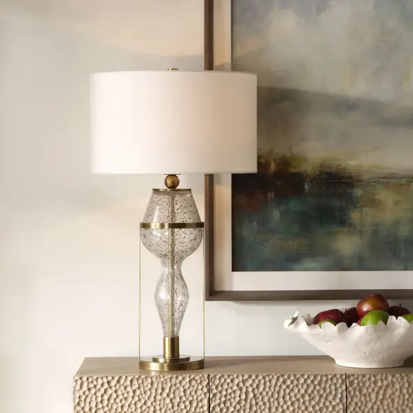Featuring A Captivating Hourglass Silhouette, This Lamp Features A Seeded Glass Base With Gray Flecks, Suspended In An Iron Support Finished In A Rich Plated Antiqued Brass. The Round Hardback Drum Shade Is Covered In A Crisp White Linen Fabric. Collection: Out Of Time Designer: Jim Parsons Finish: Antique Brass Glass: Round Hardback Drum Shade Width: 16.00" Height: 29.75" Length: 16.00" Weight: 9.50 lb Number of Bulbs: 1 Maximum Wattage: 150.00 W Bulb Type: A Voltage: 110.00 V Material: Iron/Glass/ Fabric Country of Origin: Vietnam Safety Rating: Indoor Only