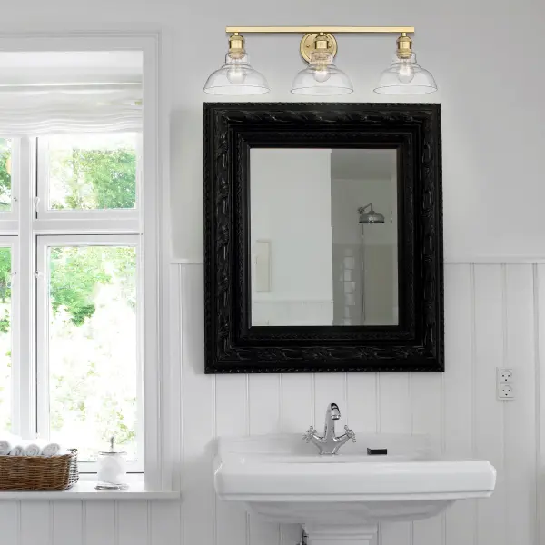 The Carver 3-Light Vanity combines contemporary elegance with industrial charm through its brushed champagne brass finish and clear glass shades. Perfect for modern bathrooms, this damp-rated fixture provides brilliant illumination while showcasing exposed bulbs for added visual interest. - alternate view