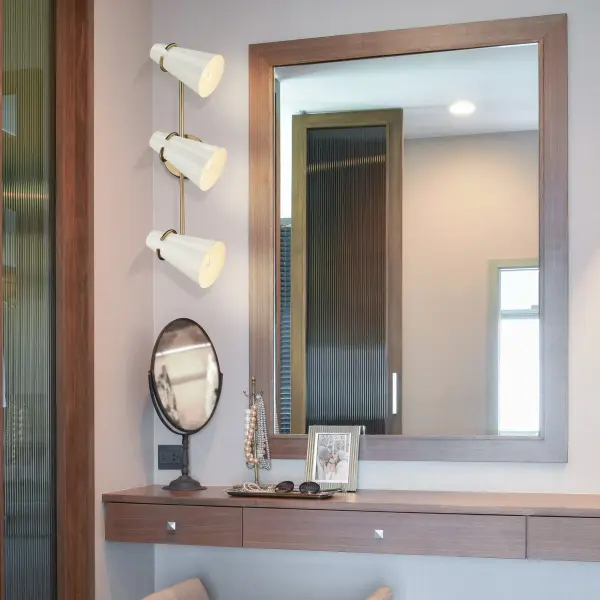 The Reeva 3-light vanity combines mid-century modern elegance with practical functionality. Its Modern Brass finish and Glossy Ecru shades create a warm, sophisticated atmosphere while adjustable heads allow customized lighting direction. - alternate view