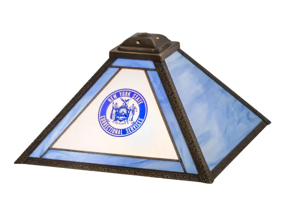 Designed with the bold New York State Correctional Services name and logo; this handsome art glass shade will enhance an office interior while featuring the artwork in Blue on a White and Blue Opal Glass background. The trim is featured in Blue art glass; while the forefront features Frosted glass; and the frame is enhanced with a Craftsman Brown finish. The shade pairs well with a base to create an appealing table lamp. The Personalized Corrections Shade is included in our American-made; American-designed; handcrafted Personalized Lighting Collection and manufactured in Yorkville; New York. This art glass shade can be ordered; and we will coordinate the custom logo with you to suit your project. Call for pricing and lead time.