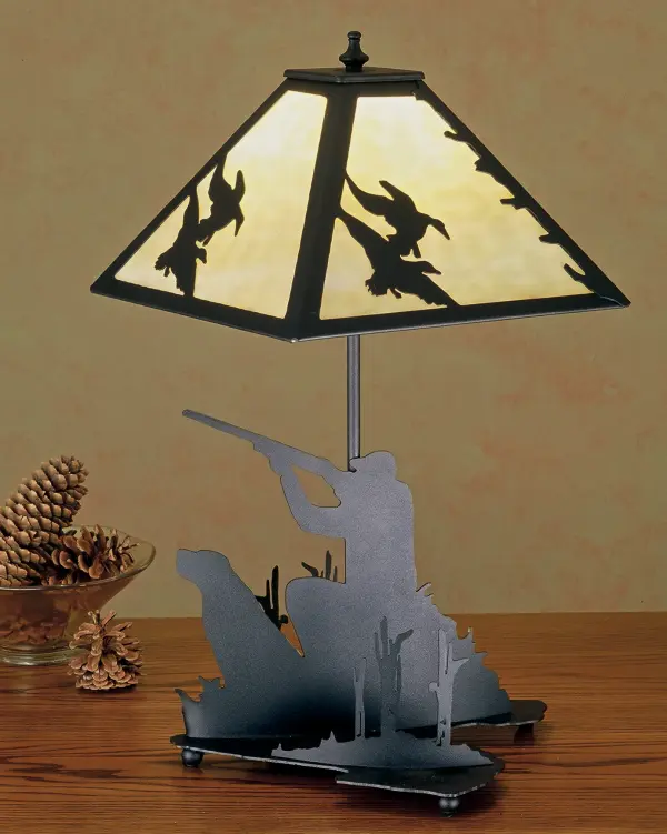 A linear square shade with ducks in flight sits above a lamp base that depicts a hunter and his retriever in a marshy blind. The table lamp has a Black finish; has Beige art glass panels and is handcrafted in the USA by Meyda artisans.