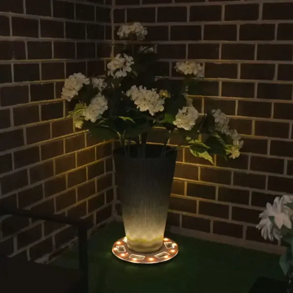 The Grand Solar Uplight Planter Base transforms ordinary planters into glowing focal points with its warm 60-lumen upward glow. This 15" wireless solar-powered base requires no wiring or electricity, charging during the day and automatically illuminating at night. Perfect for patios, porches, walkways, and poolside settings. - alternate view