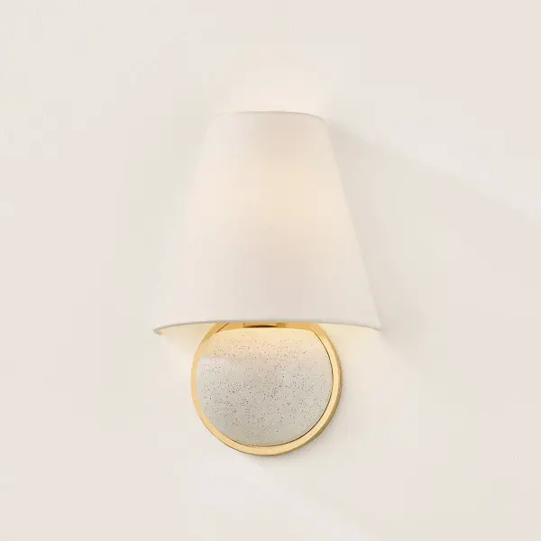 The Marlena sconce combines geometric forms with artisanal craftsmanship, featuring an aged brass racetrack arm that cradles a speckled ceramic ball beneath a white linen shade. This clever mix creates a graphic, standout fixture. - alternate view