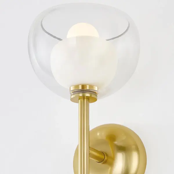 The Cortney Wall Sconce features a sophisticated double-glass design with an opal inner shade captured within clear glass, complemented by Aged Brass hardware. This trendy fixture offers beautiful light diffusion and reflection. - alternate view