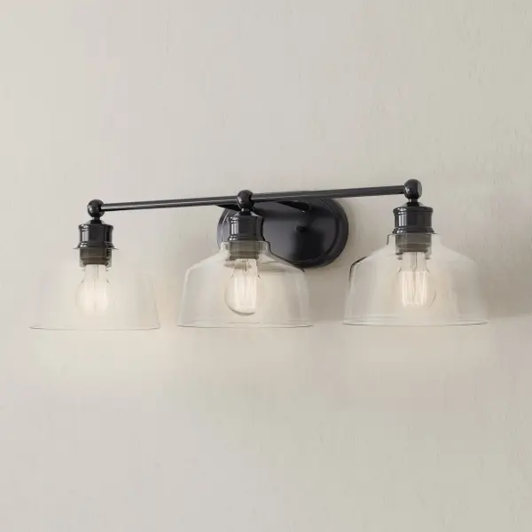 The Singleton three-light vanity fixture combines industrial style with farmhouse charm. Clear glass shades and brushed nickel finish create a versatile look that complements modern and traditional bathrooms alike. - alternate view