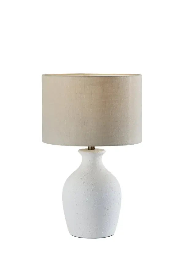 The Margot table lamp blends farmhouse and coastal aesthetics with its textured white ceramic jug-shaped base and light beige fabric drum shade. At 25" tall, it's perfectly proportioned for bedside tables and desks, casting soft, warm light through its textured shade.
