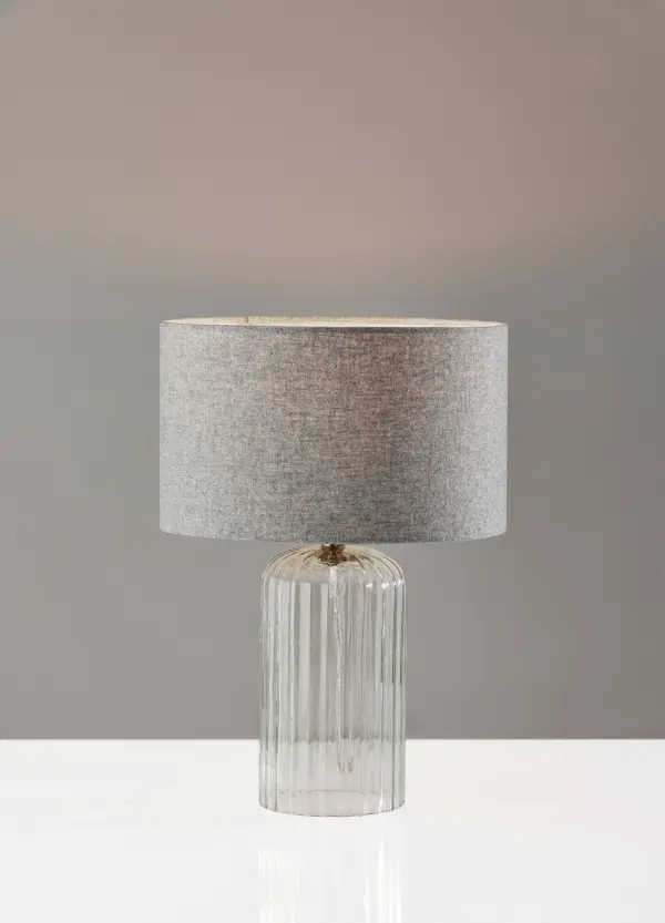 The Carrie Small Table Lamp blends coastal charm with contemporary elegance through its clear ribbed glass base and antique brass neck. The soft-touch grey drum shade provides gentle illumination perfect for bedside or desk use. - alternate view