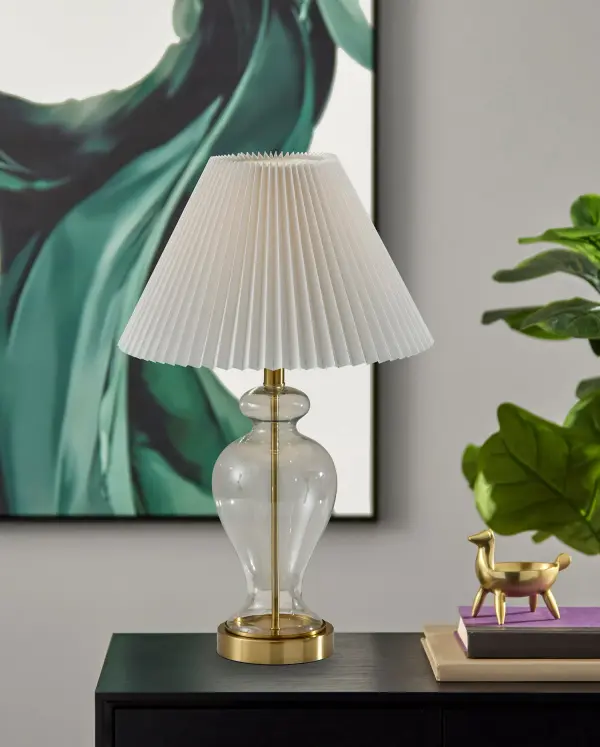 The Sonata Table Lamp blends vintage charm with modern functionality through its clear glass base and antique brass accents. The off-white pleated shade creates soft, inviting illumination perfect for bedside or desk use. - alternate view