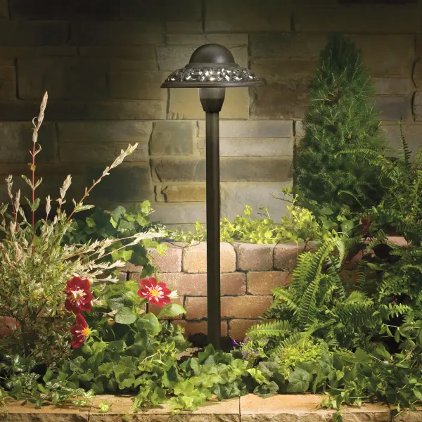 This low-voltage path light combines functional outdoor illumination with artisanal design. The cast aluminum dome features a sculpted floral band with pierced openings that create subtle light patterns along pathways and garden beds.
