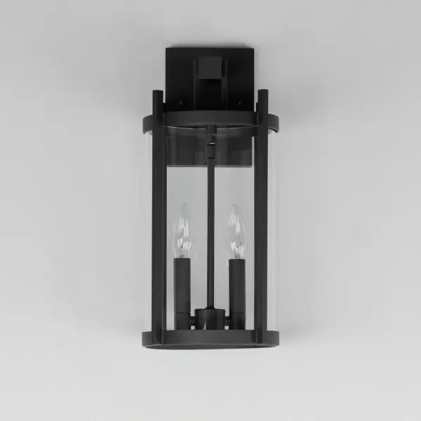 Squared columns in Matte Black finish tower above the cylindrical glass shade rated for outdoor applications. Available in a more modern clear glass option or hammered glass. A rectangular hook detail carries the square form through to the backplate. A modern interpretation of a bell tower, these outdoor pendants and sconces are a cleaned up, transitional look for exteriors. - alternate view