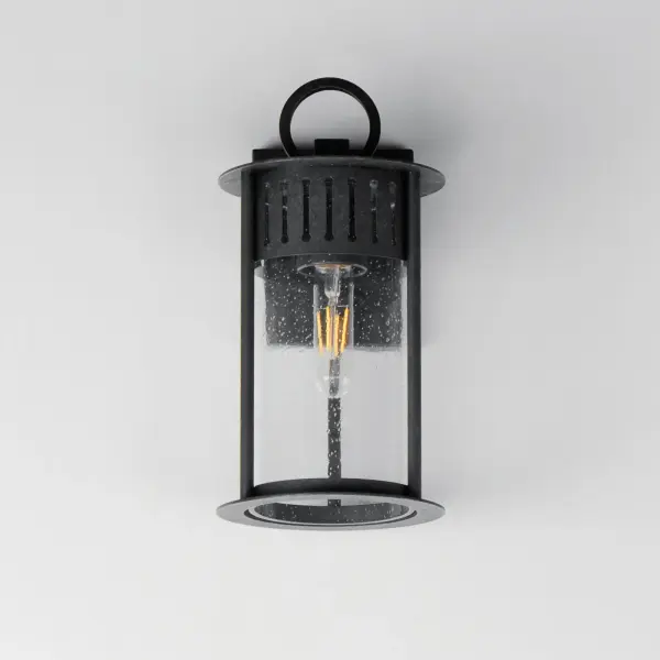 Inspired by the rounded towers of Windsor Castle, this outdoor lighting collection features a hand-applied Black Patina finish on Stainless Steel. A slotted filigree along the top of the cylinder frame allows light to escape, while the clear seeded glass obscures the light source enough to soften the glare a light bulb casts. - alternate view