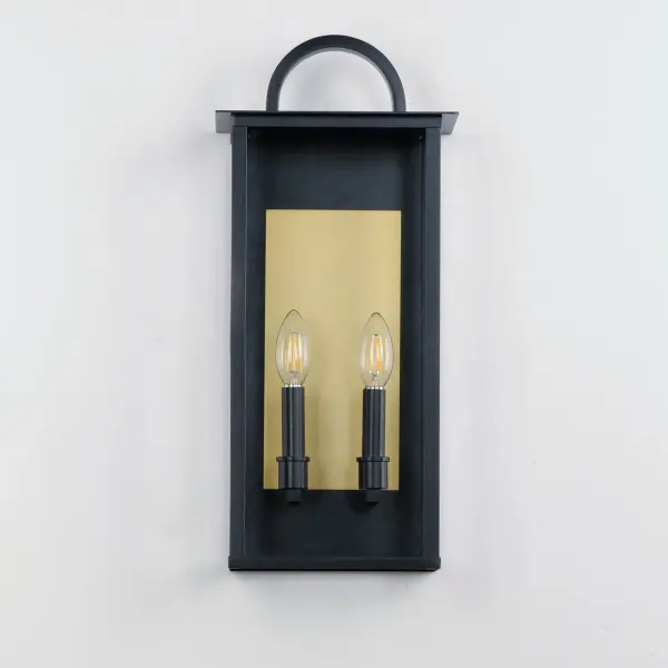 The Manchester series is inspired by classic coach lanterns and consists of minimally extended sconces in a variety of sizes that have been modified with simplified embellishments. The black frames have been modified with a squared arc handle and a contrasting Burnished Gold plate that reflects clusters of candle lights surrounded by Clear glass panels. This collection references historic exterior sconces while modernizing their appearance. Complement the design with tubular lamps to accentuate the verticality of the frames. - alternate view