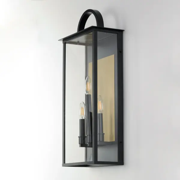 The Manchester series is inspired by classic coach lanterns and consists of minimally extended sconces in a variety of sizes that have been modified with simplified embellishments. The black frames have been modified with a squared arc handle and a contrasting Burnished Gold plate that reflects clusters of candle lights surrounded by Clear glass panels. This collection references historic exterior sconces while modernizing their appearance. Complement the design with tubular lamps to accentuate the verticality of the frames. - alternate view