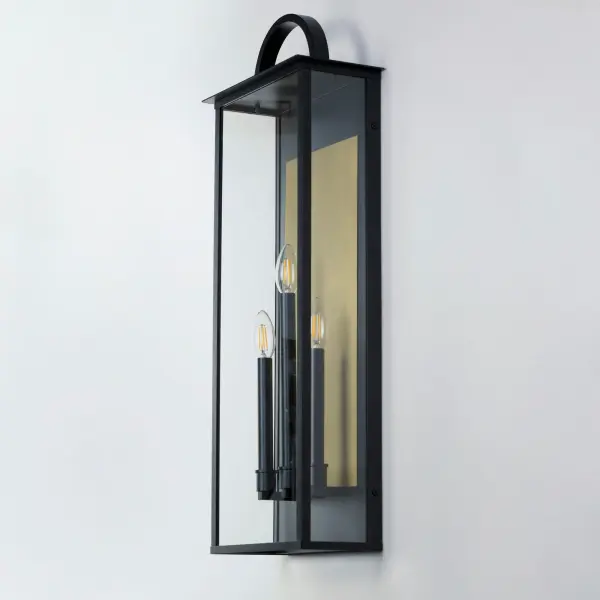 The Manchester series is inspired by classic coach lanterns and consists of minimally extended sconces in a variety of sizes that have been modified with simplified embellishments. The black frames have been modified with a squared arc handle and a contrasting Burnished Gold plate that reflects clusters of candle lights surrounded by Clear glass panels. This collection references historic exterior sconces while modernizing their appearance. Complement the design with tubular lamps to accentuate the verticality of the frames. - alternate view