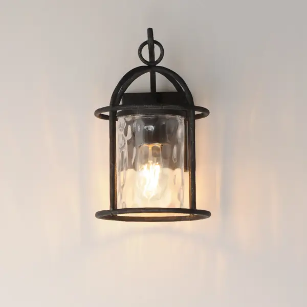 Inspired by classic European lanterns and handcrafted ironwork, the Del Mar collection brings timeless sophistication to exterior spaces. Its airy open-frame silhouette is crafted of hammered iron, elegantly arched with a graceful hook detail that nods to historic gas lamps. Inside the frame, a delicate Water glass cylinder shimmers with texture, diffusing light in a soft, atmospheric glow. Designed for lasting beauty in the elements, each piece is finished in a richly layered Blacksmith finish--a deep near-black tone highlighted by silvery brushstrokes, adding artisanal character to the silhouette. Whether flanking a grand entry or lining a garden path, Del Mar offers a refined approach to outdoor lighting with enduring presence. - alternate view