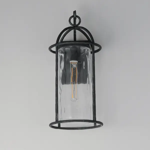 Inspired by classic European lanterns and handcrafted ironwork, the Del Mar collection brings timeless sophistication to exterior spaces. Its airy open-frame silhouette is crafted of hammered iron, elegantly arched with a graceful hook detail that nods to historic gas lamps. Inside the frame, a delicate Water glass cylinder shimmers with texture, diffusing light in a soft, atmospheric glow. Designed for lasting beauty in the elements, each piece is finished in a richly layered Blacksmith finish--a deep near-black tone highlighted by silvery brushstrokes, adding artisanal character to the silhouette. Whether flanking a grand entry or lining a garden path, Del Mar offers a refined approach to outdoor lighting with enduring presence. - alternate view