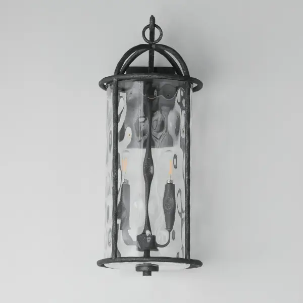 Inspired by classic European lanterns and handcrafted ironwork, the Del Mar collection brings timeless sophistication to exterior spaces. Its airy open-frame silhouette is crafted of hammered iron, elegantly arched with a graceful hook detail that nods to historic gas lamps. Inside the frame, a delicate Water glass cylinder shimmers with texture, diffusing light in a soft, atmospheric glow. Designed for lasting beauty in the elements, each piece is finished in a richly layered Blacksmith finish--a deep near-black tone highlighted by silvery brushstrokes, adding artisanal character to the silhouette. Whether flanking a grand entry or lining a garden path, Del Mar offers a refined approach to outdoor lighting with enduring presence. - alternate view