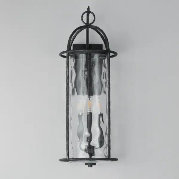 Inspired by classic European lanterns and handcrafted ironwork, the Del Mar collection brings timeless sophistication to exterior spaces. Its airy open-frame silhouette is crafted of hammered iron, elegantly arched with a graceful hook detail that nods to historic gas lamps. Inside the frame, a delicate Water glass cylinder shimmers with texture, diffusing light in a soft, atmospheric glow. Designed for lasting beauty in the elements, each piece is finished in a richly layered Blacksmith finish--a deep near-black tone highlighted by silvery brushstrokes, adding artisanal character to the silhouette. Whether flanking a grand entry or lining a garden path, Del Mar offers a refined approach to outdoor lighting with enduring presence. - alternate view