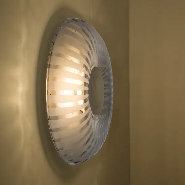 The Spira sconce features a distinctive toroid-shaped blown smoke glass shade with radially patterned frosted stripes. Its indirect LED lighting beautifully highlights the etched design, creating ambient illumination perfect for contemporary spaces.