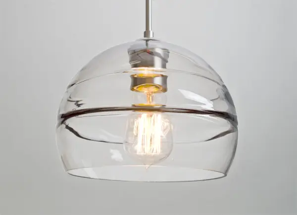 The Spirit 8 Clear Pendant features a distinctive circular optical pattern blown into the interior wall, creating dramatic light play and visual interest. The clear glass showcases the lamp filament while the sculpted layer projects beautiful patterns onto surrounding surfaces. - alternate view